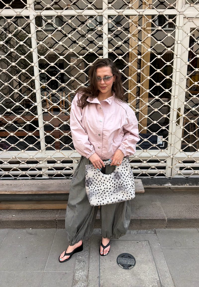 Femme portant une veste rose, un pantalon ample à carreaux, des sandales noires, tenant un sac à main à pois, debout devant une grille métallique.