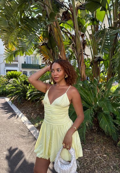 Vestido amarillo claro sin mangas con escote en V, cuerpo fruncido y falda acampanada. Accesorios incluyen un bolso blanco y joyas doradas.