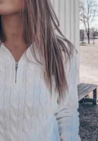 Weißer Kabelstrickpullover mit einem Viertel-Reißverschluss-Kragen, der mit strukturierten Mustern und einer lockeren Passform versehen ist, vor einer natürlichen Außenkulisse.