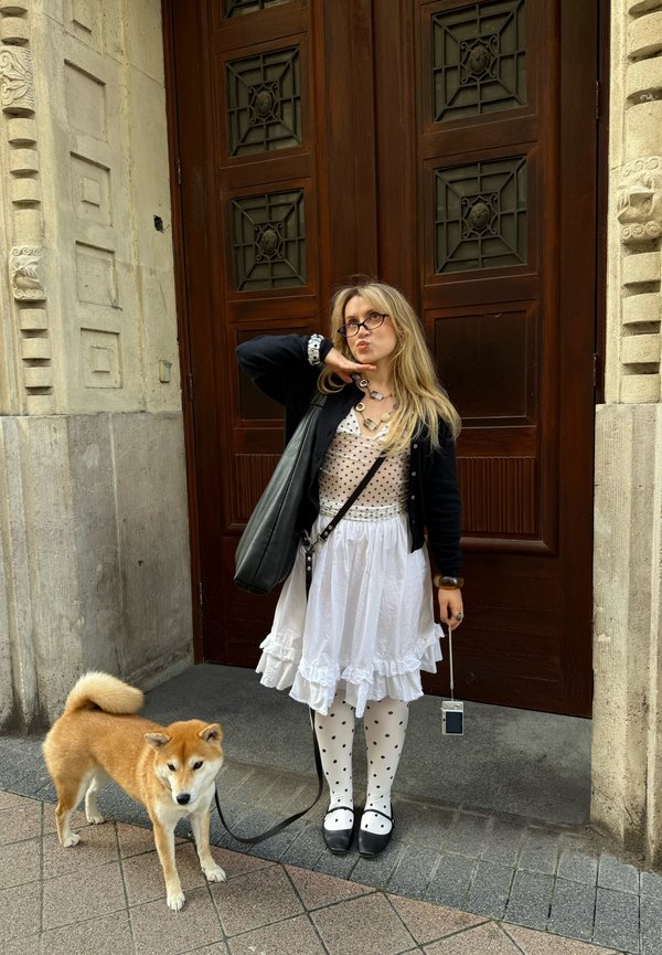 Uma pessoa vestida com um cardigã preto e um vestido branco com babados está ao lado de um cão Shiba Inu num passeio pavimentado. O vestido tem detalhes de bolinhas.