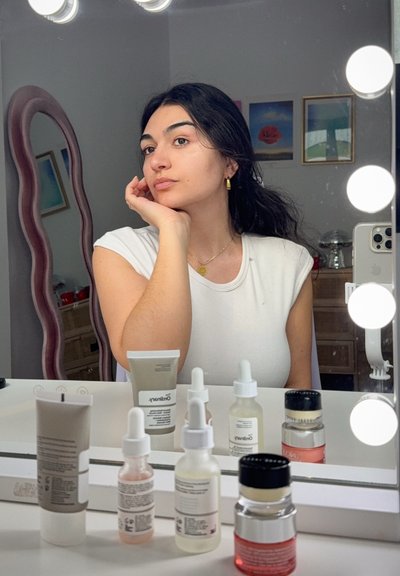 Joven mujer con cabello oscuro y camisa blanca se sienta en el tocador con productos para el cuidado de la piel, mirando en un espejo rodeado de luces redondas.