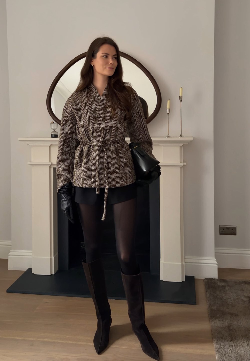 Femme portant une veste ceinturée à motifs, une jupe noire, des collants, des bottes hautes et des gants, debout à l'intérieur près d'une cheminée avec un miroir décoratif.