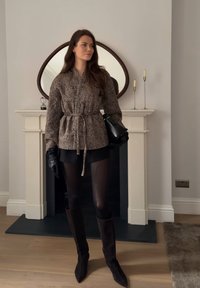 Femme portant une veste ceinturée à motifs, une jupe noire, des collants, des bottes hautes et des gants, debout à l'intérieur près d'une cheminée avec un miroir décoratif.