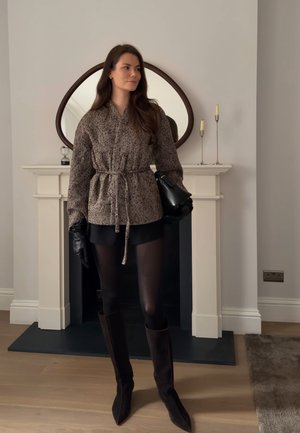 Mujer con chaqueta estampada con cinturón, falda negra, medias, botas hasta la rodilla y guantes, de pie en el interior cerca de una chimenea con un espejo decorativo.