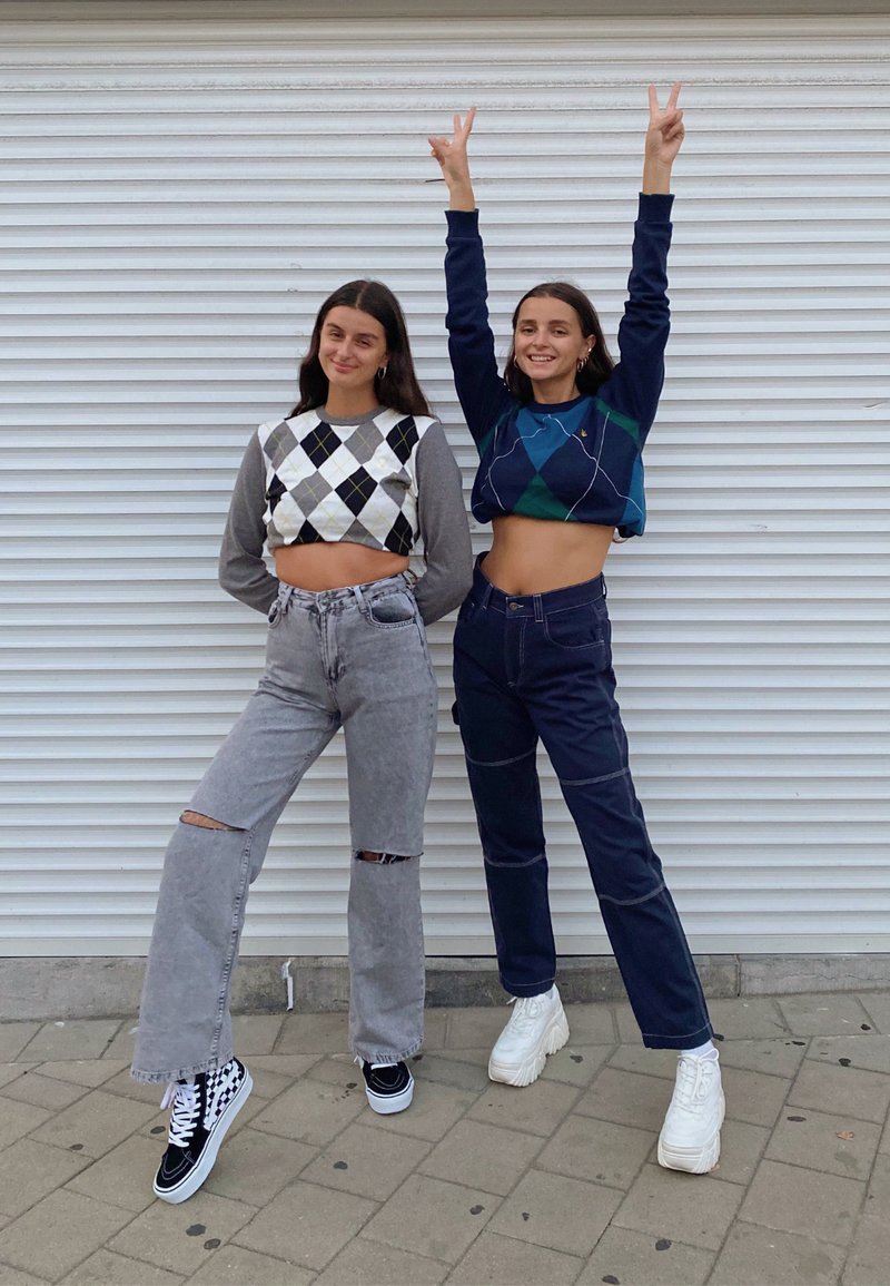 To modeller står og poserer. Den ene har en kortærmet argyle sweater i grå og sort på, kombineret med høj talje grå jeans. Den anden har en navy kortærmet sweater og mørke jeans.