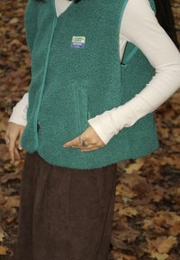 Włóczkowa kamizelka w kolorze turkusowym z zamkiem i kieszeniami bocznymi, noszona na białej bluzce z długim rękawem. Brąskowa spódnica o strukturze, ręka spoczywa na kamizelce.