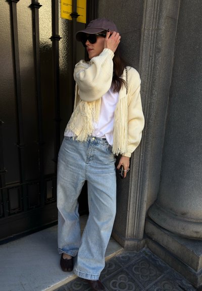 Mujer con jeans claros de gran tamaño, camiseta blanca, suéter crema con flecos, gorra negra y gafas de sol, apoyada contra un pilar de piedra cerca de una puerta negra.