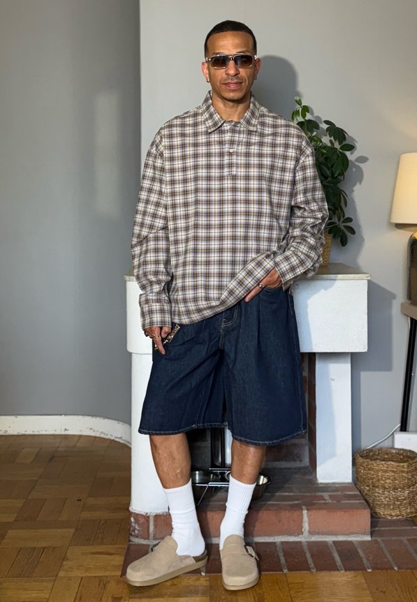 Mann mit Sonnenbrille, kariertem Hemd, Jeansshorts, weißen Socken und beigen Slip-On-Schuhen steht drinnen nahe einem Kamin und einer Pflanze.