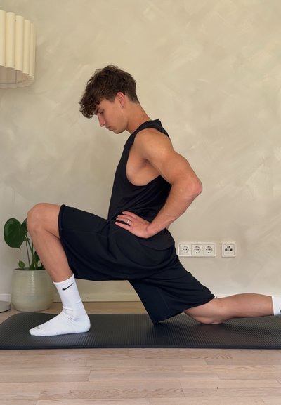 Joven con ropa deportiva negra realiza una zancada en una esterilla de yoga en interior, estirando sus flexores de cadera, con las manos en las caderas y la cabeza ligeramente inclinada.