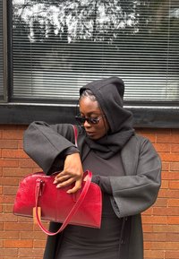 Borsa a mano in pelle rossa con una forma strutturata e una texture liscia. Presenta doppi manici e una cerniera superiore, tenuta da una persona con un cappotto nero.