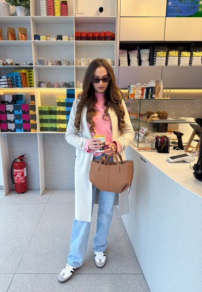 Trinchera beige claro sobre un suéter rosa, jeans azules y zapatillas grises. Sostiene una taza a rayas y un bolso beige. Entorno de café moderno.