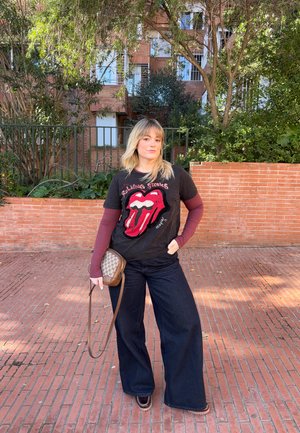 Joven con cabello rubio que lleva una camiseta de los Rolling Stones, pantalones anchos oscuros, y sostiene un pequeño bolso marrón cruzado al aire libre.