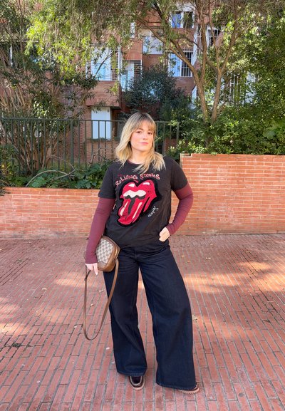 Joven con cabello rubio que lleva una camiseta de los Rolling Stones, pantalones anchos oscuros, y sostiene un pequeño bolso marrón cruzado al aire libre.