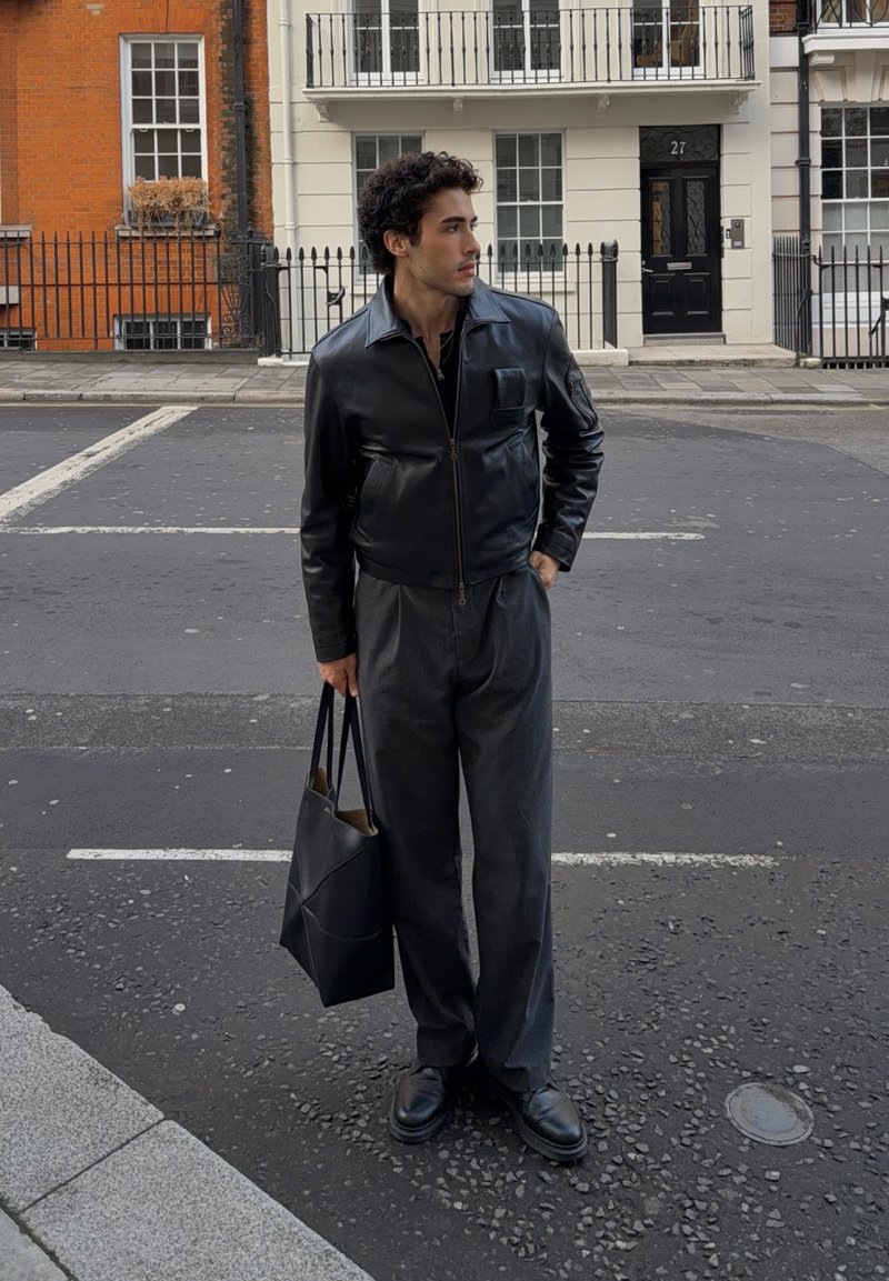 Jeune homme debout dans la rue portant une veste en cuir noire, un pantalon gris, des chaussures noires, tenant un sac fourre-tout noir, avec des bâtiments urbains en arrière-plan.