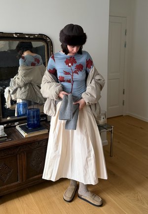 Persona con chaqueta acolchada beige, blusa azul con flores, falda crema, sombrero marrón, zapatillas beige, sosteniendo guantes de punto grises en una habitación con suelo de madera.