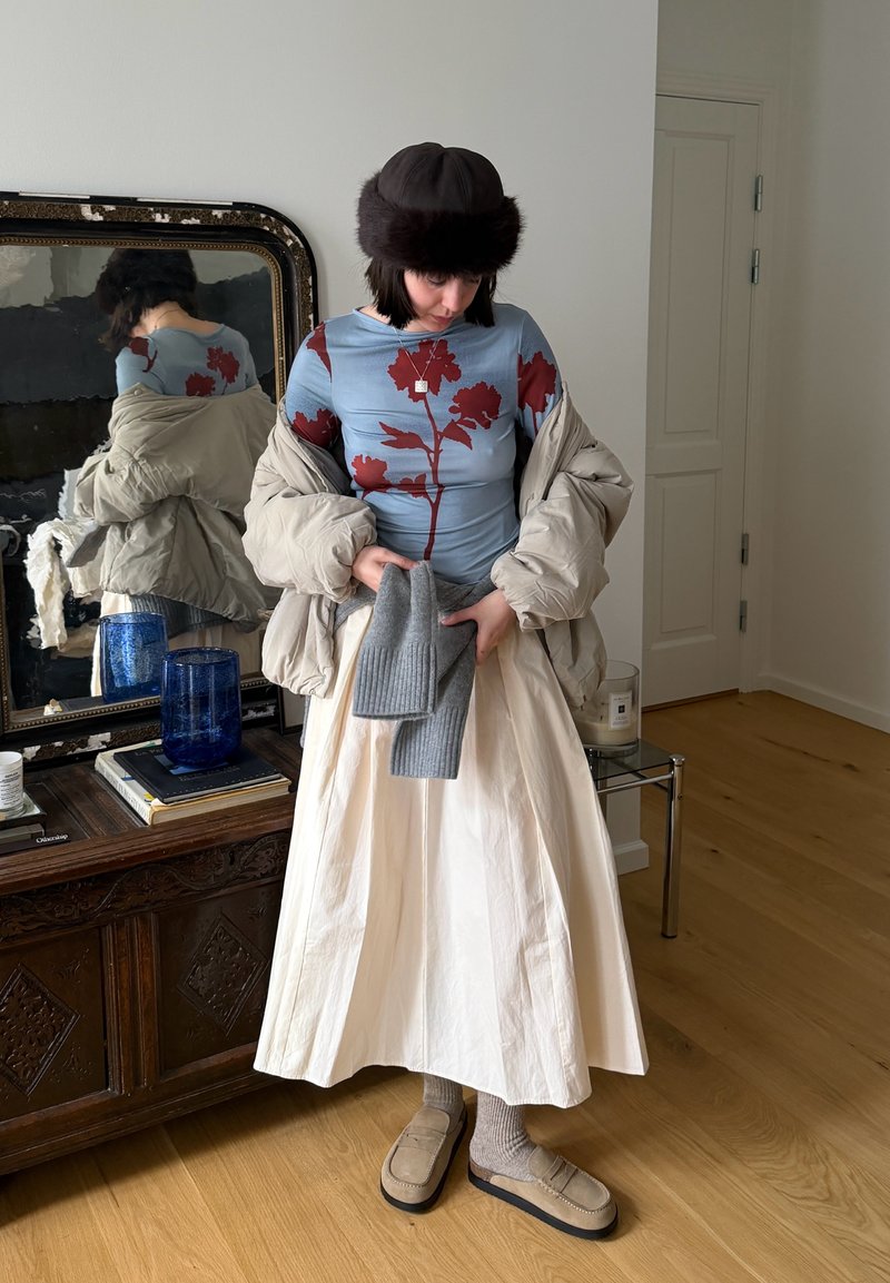 Personne portant une doudoune beige, un haut bleu à fleurs, une jupe crème, un chapeau marron, des chaussons beiges, tenant des gants en tricot gris dans une pièce avec un sol en bois.