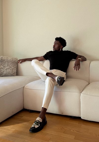 Hombre con pendientes y anillos, vestido con camisa negra y pantalones blancos, sentado con las piernas cruzadas en un sofá beige en una habitación con suelo de madera.