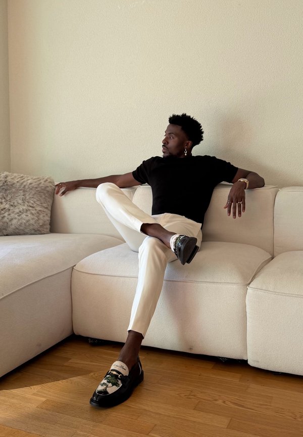 Hombre con pendientes y anillos, vestido con camisa negra y pantalones blancos, sentado con las piernas cruzadas en un sofá beige en una habitación con suelo de madera.