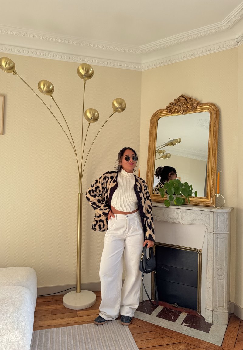 Femme portant des lunettes de soleil, une veste imprimée léopard, un col roulé blanc, un pantalon blanc large, debout à côté d'une lampe de sol dorée et d'une cheminée ornée avec miroir.