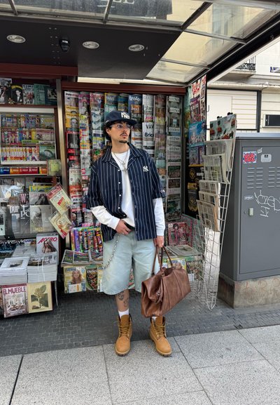 Un hombre está de pie frente a un quiosco de prensa, vistiendo una camisa de rayas de manga corta, pantalones cortos de mezclilla claros, botas marrones y sosteniendo un bolso de cuero marrón.