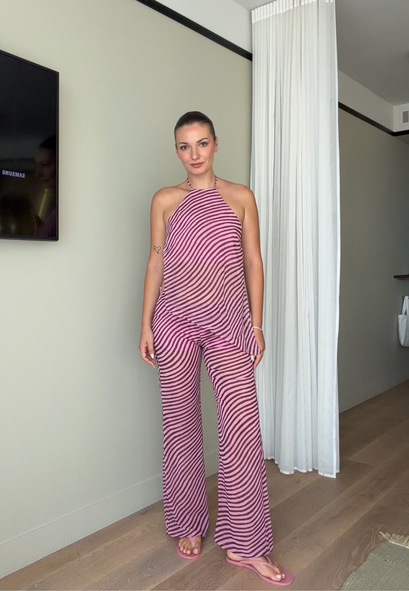 Vrouw staat binnen, draagt een roze en paarse gestreepte haltertop en bijpassende wijde broek met roze teenslippers.