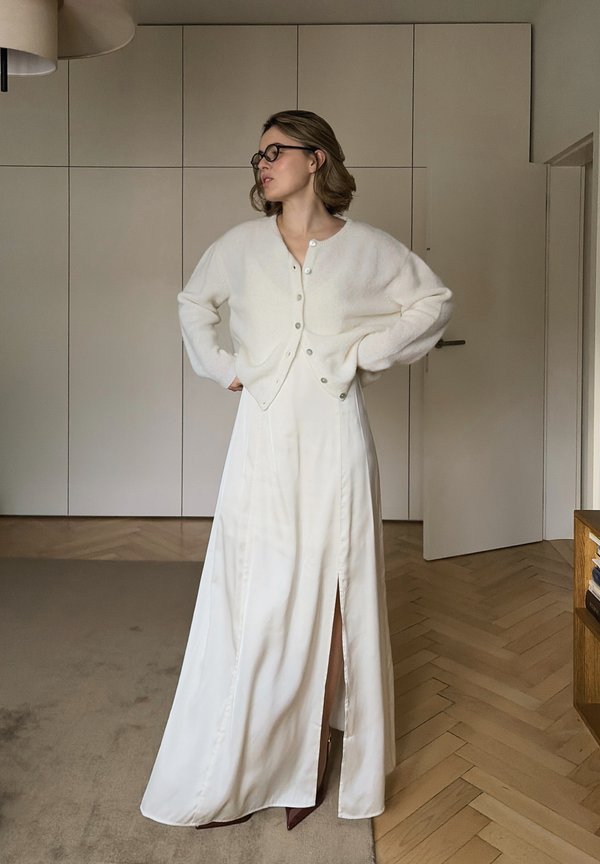 Witte oversized cardigan met knopen, gecombineerd met een lange witte rok met een zij-split, tegen een minimalistisch interieur.
