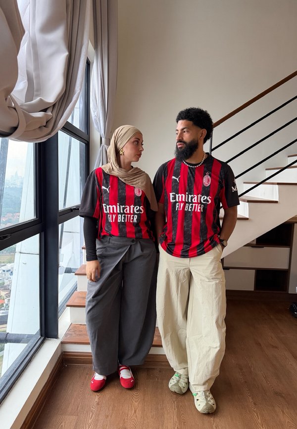 Un hombre y una mujer con camisetas a juego del AC Milan están de pie uno al lado del otro junto a una ventana y una escalera, mirándose mutuamente.