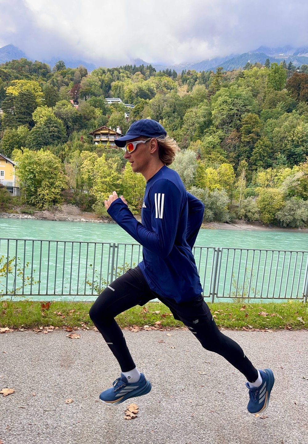 En løper i en blå langermet skjorte og svarte tights jogger langs en sti ved en elv, iført caps og solbriller.