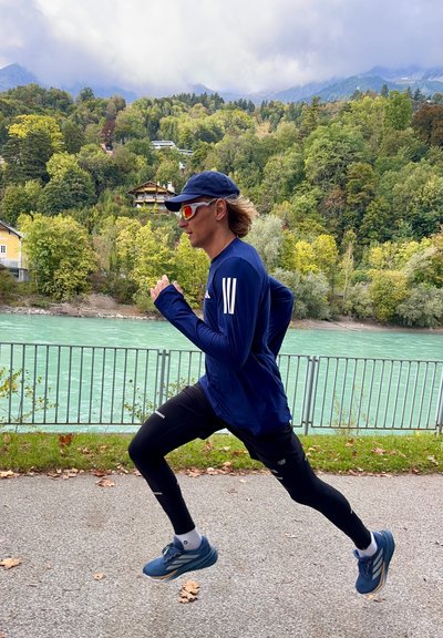 Un corredor con una camiseta de manga larga azul y leggings negros corre por un sendero cerca de un río, llevando una gorra y gafas de sol.
