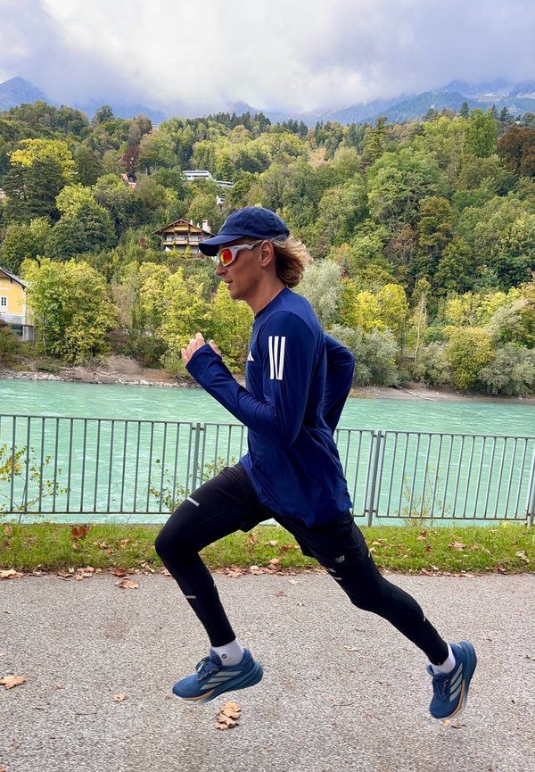 En løper i en blå langermet skjorte og svarte tights jogger langs en sti ved en elv, iført caps og solbriller.