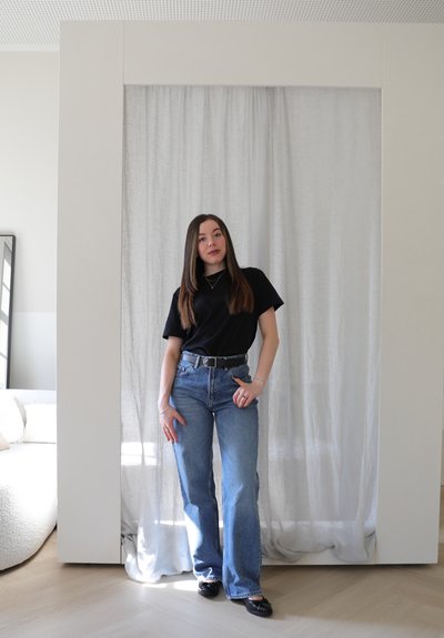 Camiseta negra, jeans azul de cintura alta con pierna recta y zapatos planos negros. Accesorios plateados complementan el atuendo casual en una habitación bien iluminada.
