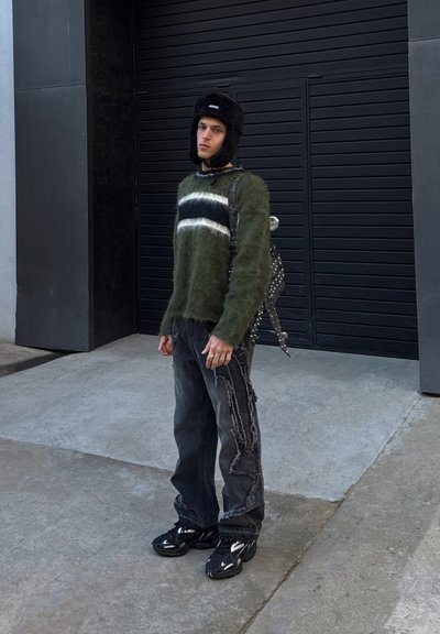 Suéter verde oliva de lana con franjas blancas, sombrero de piel sintética negra, jeans negros desgastados y zapatillas negras con detalles blancos, de pie contra una pared oscura.