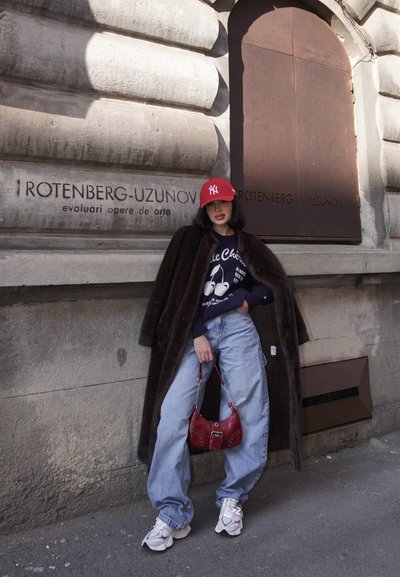 Abrigo de piel sintética marrón, sudadera gráfica azul marino, vaqueros ligeros sueltos de color azul claro, zapatillas blancas y un bolso rojo con detalle de cadena; posando frente a una pared texturizada.