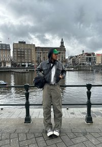 A person stands by a river wearing a black leather jacket, grey oversized trousers, and a green cap, with a black messenger bag.