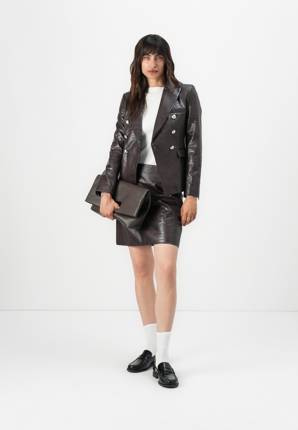 Hnědý, texturovaný kožený blazer s dvojitým stříbrným zapínáním, v kombinaci s odpovídající sukní a černými mokasíny. Drží tmavou kabelku.