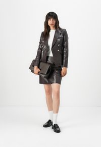 Hnědý, texturovaný kožený blazer s dvojitým stříbrným zapínáním, v kombinaci s odpovídající sukní a černými mokasíny. Drží tmavou kabelku.