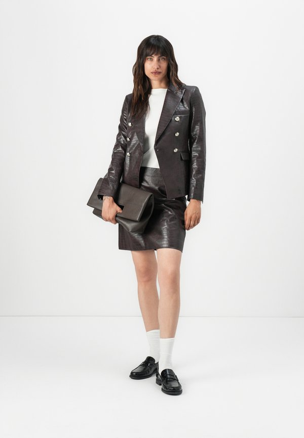 Blazer in pelle marrone strutturata, con bottoni argento doppi, abbinato a una gonna coordinata e mocassini neri. Tiene una pochette scura.