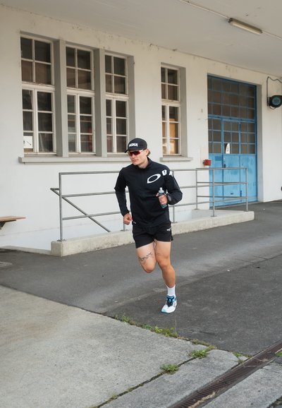 Corredor con una camiseta de manga larga negra y pantalones cortos, gorra negra y gafas de sol, sosteniendo una botella de agua, con tatuajes visibles en las piernas.