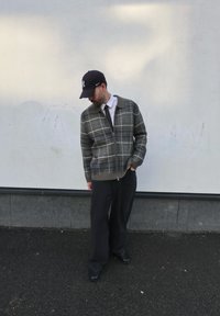 Pull zippé à carreaux gris et verts avec col, associé à un pantalon noir à rayures fines et une casquette sombre. Debout sur une surface texturée.
