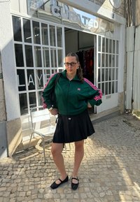 Green zip-up jacket with pink accents, black pleated skirt, and black ballerina flats; styled with glasses and a bag; cobblestone background.