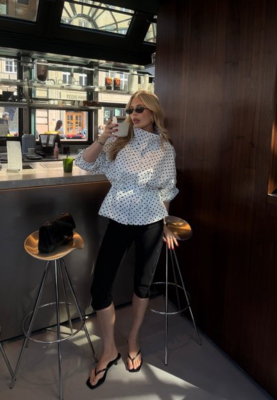 Mujer con blusa blanca de lunares y pantalones capri negros bebe café en el mostrador de la cafetería con dos taburetes y un bolso negro.