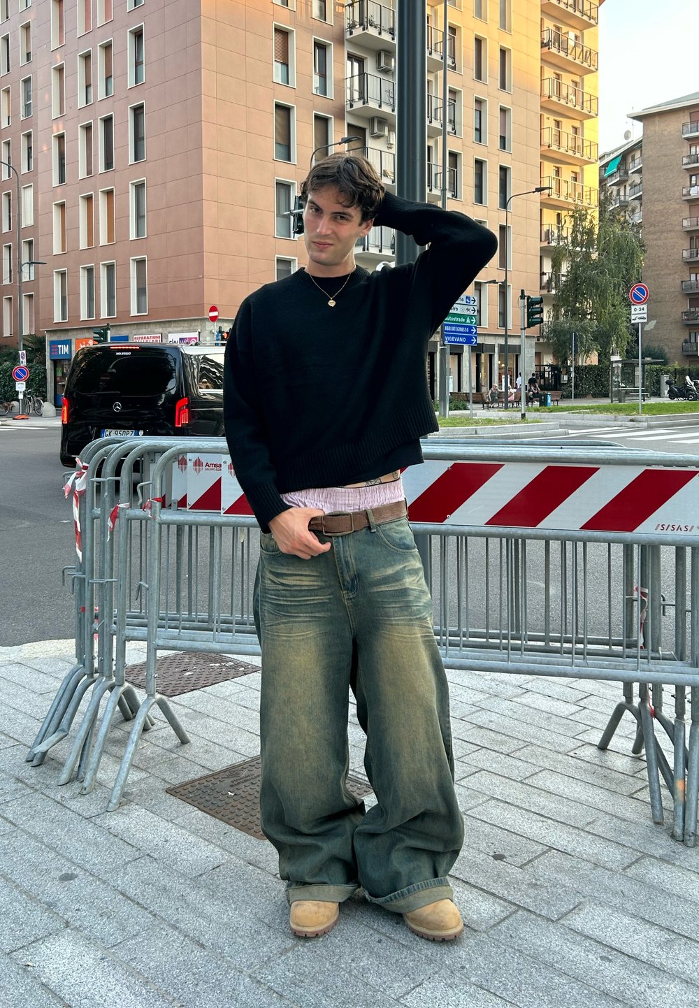 Ung mand i mørk sweater og brede jeans står på en urban fortov nær metalbarrierer og gadeskilt foran lejlighedsbygninger.