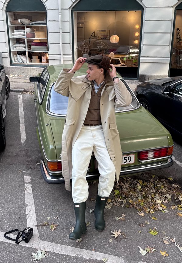 Jeune homme ajustant son chapeau tout en étant assis sur le capot d'une voiture vintage verte garée dans une rue de la ville à côté d'une vitrine.