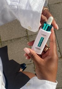 Sérum L'Oréal Paris dans un flacon en verre blanc avec un bouchon turquoise. L'étiquette indique "Bright Reveal Niacinamide." Bouchon à goutte pour l'application.