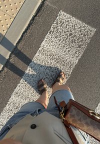 Brune sandaler med remmer, lyseblå denim shorts med frynser, grå skjorte, og en brun crossover-veske på et teksturert hvitt fotgjengerfelt.