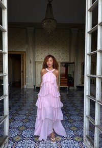 Vestido de color rosa claro, de capas y con diseño de halter, que presenta capas con volantes y un detalle de nudo en el pecho. Textura suave, forma fluida.