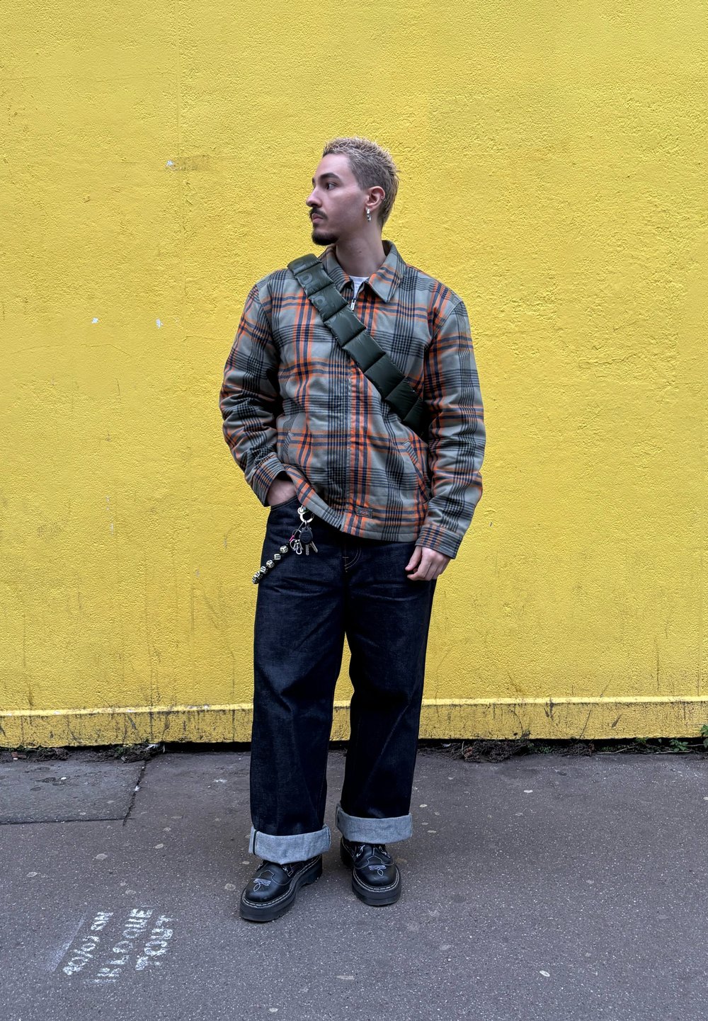 Veste à carreaux avec des tons gris, orange et noir ; jean noir ample avec les revers roulés ; sac bandoulière vert ; chaussures noires avec des accents argentés.