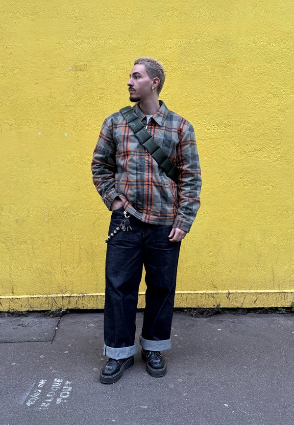 Jachetă în carouri cu tonuri de gri, portocaliu și negru; blugi negri largi cu tivuri rulate; geantă verde purtată transversal; pantofi negri cu accente argintii.