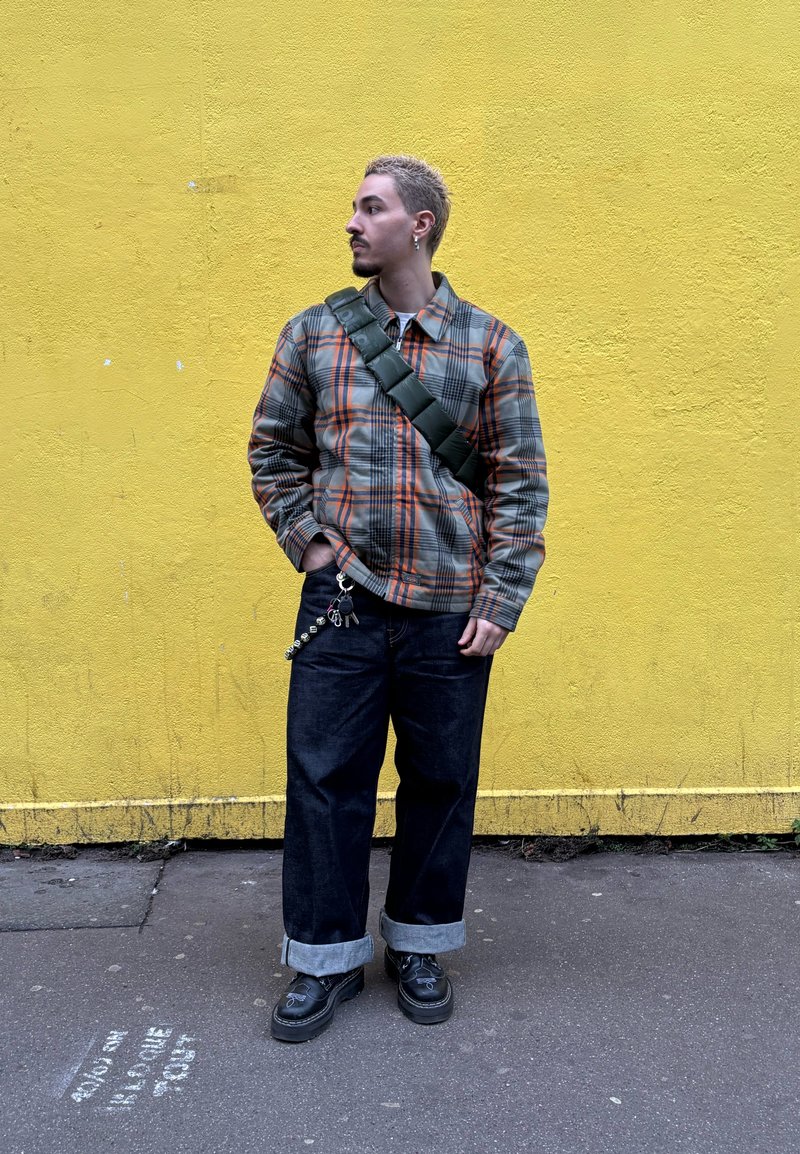 Veste à carreaux aux tons gris, orange et noir ; jean noir ample avec revers roulés ; sac bandoulière vert ; chaussures noires avec accents argentés.