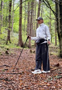 Osoba w białej kurtce i czarnych spodniach stoi na leśnej ścieżce, trzymając kijki trekkingowe, otoczona liściastymi drzewami i jesiennymi liśćmi.